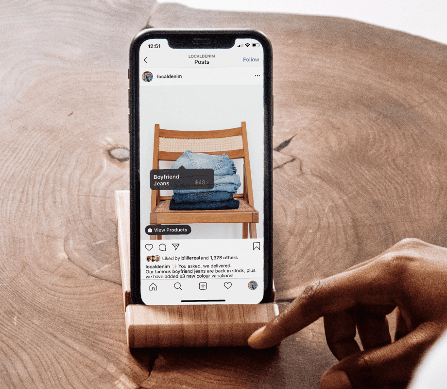 Phone standing upright in a dark brown wooden phone holder. Onscreen is a photo of three pairs of boyfriend jeans of various blue shades folded on a light brown wooded chair. 